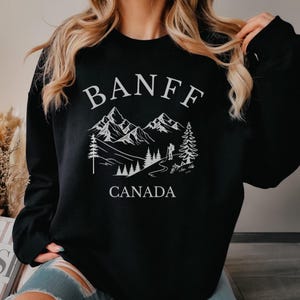 May include: A black long-sleeve sweatshirt featuring a white graphic print. The design displays the text BANFF arched over a mountain range, pine trees, and a hiker silhouette, with the word CANADA printed below the landscape illustration.