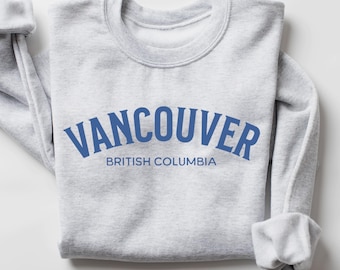 May include: A light gray crewneck sweatshirt featuring the blue text VANCOUVER in an arched font, with the words BRITISH COLUMBIA printed in a smaller, straight blue font directly underneath the city name on the front of the garment.