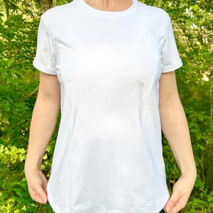 Pode incluir: Uma camiseta esportiva branca de manga curta. A camiseta com gola redonda tem um design ajustado com costuras que contornam o corpo. O tecido parece ser um material leve e respirável, adequado para exercícios ou uso casual.