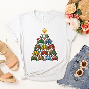 May include: A white short-sleeved t-shirt featuring a graphic print of a Christmas tree constructed from various colorful video game controllers. The tree is topped with a yellow star and decorated with small ornaments and string lights.