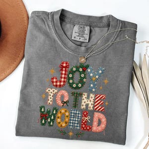 May include: A charcoal gray short-sleeve t-shirt featuring a graphic print that reads JOY TO THE WORLD. The letters are designed with various festive patterns including red plaid, green floral, blue stars, and holiday motifs like bows and holly.