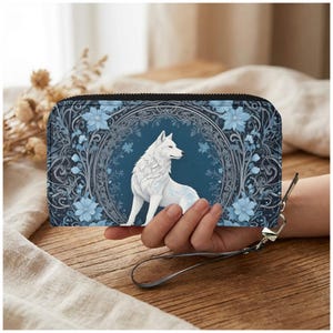 May include: A blue and black wallet featuring a white wolf design. The wallet has a zipper closure and a wrist strap. The wolf is surrounded by a floral and ornamental design in shades of blue and gray.