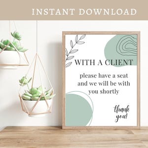 May include: Printable sign with a green, white, and black color scheme. The sign reads "With a Client, please have a seat and we will be with you shortly, thank you!"