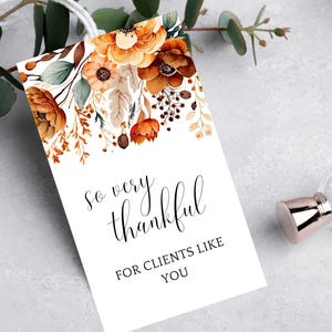 Op de afbeelding: Een witte kaart met een bloemenontwerp in tinten oranje, bruin en groen. De tekst op de kaart luidt "So very thankful for clients like you."