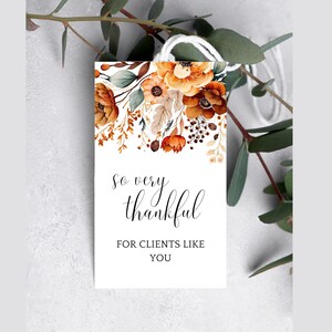 Op de afbeelding: Een wit label met een bloemenontwerp in tinten oranje, bruin en cr&egrave;me. De tekst "So very thankful for clients like you" is in zwart gedrukt.