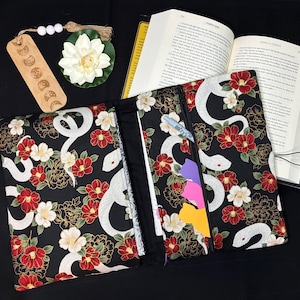 May include: A fabric book cover featuring a black background with a pattern of white snakes, red and white flowers, and gold outlines. The cover includes an internal pocket containing a pen and colorful sticky notes, positioned next to an open book.