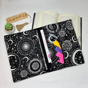 May include: A black fabric book sleeve featuring a white celestial pattern of moons, suns, and stars. The sleeve is open, revealing a book inside, a light blue pen, and three cat-shaped sticky notes in yellow, purple, and pink. A wooden moon phase bookmark with a beige tassel and a small potted succulent are nearby.