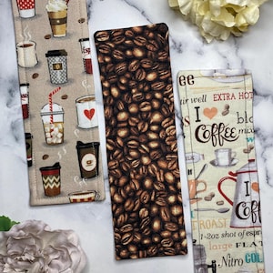 May include: Three rectangular fabric bookmarks featuring coffee-themed designs. The left bookmark has various coffee cups on a beige background. The center bookmark displays a pattern of dark roasted coffee beans. The right bookmark features text and coffee illustrations on a cream background.