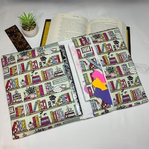 May include: A fabric book cover featuring a repeating pattern of colorful bookshelves with various books, potted plants, and sleeping cats. The cover is open, revealing a white interior lining, a grey pen, and several cat-shaped sticky notes.