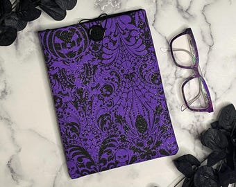 Pode incluir: Uma capa de livro roxa e preta com tema de Halloween com fecho de botão. A capa apresenta um padrão de caveiras, teias de aranha e outros desenhos assustadores. Um par de óculos com armação roxa repousa ao lado da capa do livro.
