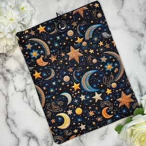 May include: A dark blue fabric book cover with a celestial design featuring gold and blue stars, moons, and crescent shapes. The cover has a button closure and is surrounded by white flowers.