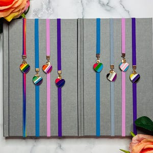 May include: Two gray journals with colorful ribbon bookmarks. Each bookmark has a heart-shaped charm with a different LGBTQ+ pride flag design. The ribbons are in shades of blue, pink, and purple. The image is decorated with roses.