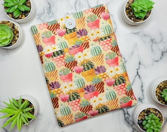 May include: A beige fabric case with a button closure, patterned with potted cacti, flowers, and succulents in various colors. The case is surrounded by small potted succulents.
