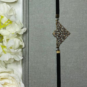 May include: A gray journal with a textured cover and a black elastic band closure. A decorative bronze-colored filigree charm and tassel are attached to the band. White flowers are in the background.
