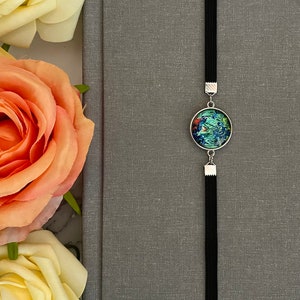 May include: A black elastic bookmark with a round, silver-toned charm featuring a colorful floral design. The bookmark is attached to a gray notebook.