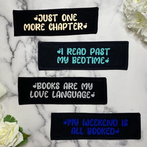 May include: Four rectangular black fabric bookmarks featuring text in different colors. The top reads JUST ONE MORE CHAPTER in yellow, the second I READ PAST MY BEDTIME in teal, the third BOOKS ARE MY LOVE LANGUAGE in white, and the bottom MY WEEKEND IS ALL BOOKED in blue.