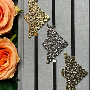 May include: Three decorative metal bookmarks with intricate designs in gold and silver, attached to black elastic bands. The bookmarks are displayed on a gray textured surface.