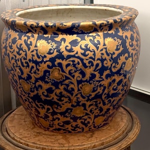May include: A large, round ceramic planter featuring a deep cobalt blue base colour with an intricate, repeating gold-toned scrolling vine and leaf pattern covering the exterior surface. The interior of the planter is a plain, off-white colour.