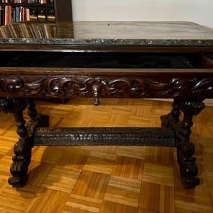 May include: An antique dark brown table with a marble top and a drawer. The table features ornate carvings on the legs and drawer front. The drawer is open, revealing a black interior. The table is on a parquet floor.
