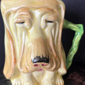 Peut inclure: Mug en céramique en forme de visage de Basset Hound. La tasse est principalement jaune avec des détails bruns, des narines noires et une bouche rose. La poignée est verte et en forme de vigne.