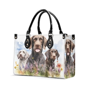 May include: White tote bag featuring a watercolor painting of several dogs in a field of flowers. The bag has black handles and accents. The dogs are various shades of brown and gray, with a blue sky background.