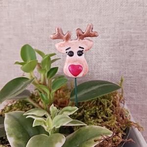 May include: A decorative reindeer plant stake with a red heart and brown antlers. The reindeer is light pink with black eyes and is placed in a pot with green plants and moss. A festive accent for indoor plants.
