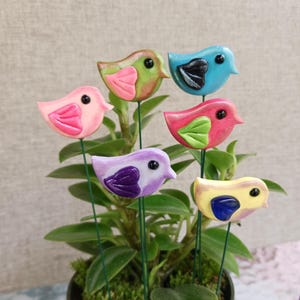 May include: A collection of six colorful bird garden stakes. Each bird is a different color, including pink, green, blue, red, purple, and yellow. The birds are attached to green wire stems and are placed in a potted plant.