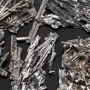 May include: Close-up of several silver-colored mineral specimens with a crystalline structure. The minerals have a metallic sheen and are arranged against a dark, textured background. The crystals are in various shapes.