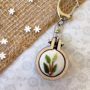 May include: A small wooden embroidery hoop keychain with a green leaf design. The hoop is attached to a silver metal key ring.
