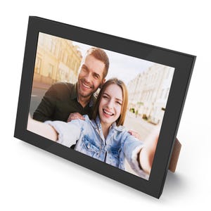 May include: A black picture frame with a wood kickstand holds a photo of a couple taking a selfie. The couple is smiling and appears to be in a city setting.