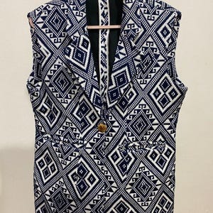 May include: Sleeveless blazer with a geometric pattern in navy blue and white. The blazer has a single wooden button closure and two front pockets. The pattern is a repeating design of diamonds and other shapes.