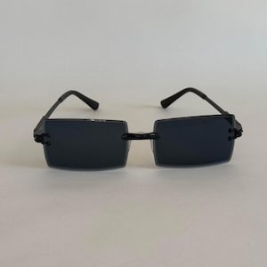 Könnte beinhalten: Rechteckige Sonnenbrille mit dunkel getönten Gläsern und einem schwarzen Rahmen. Die Sonnenbrille hat ein minimalistisches Design mit einem dünnen Rahmen und schlanken Bügeln. Die Gläser sind dunkelgrau.