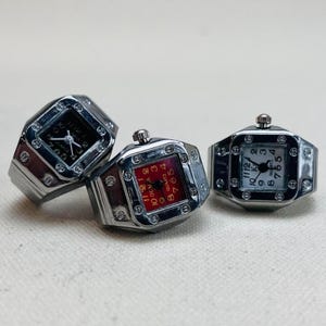 Puede incluir: Tres relojes de anillo plateados con caras cuadradas. El primer reloj tiene una cara negra con números y manecillas blancas. El segundo reloj tiene una cara roja con números y manecillas blancas. El tercer reloj tiene una cara blanca con números y manecillas negras.