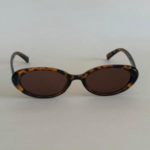 May include: Oval-shaped sunglasses with a tortoiseshell pattern frame and dark brown lenses. The sunglasses have a classic design, suitable for various face shapes. The frame features a mix of brown and black hues.