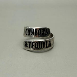 May include: A silver-colored wrap ring with the words "COWBOYS & TEQUILA" engraved in black. The ring has a textured, slightly tarnished appearance, and the text is in a bold, sans-serif font.
