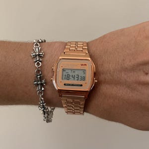 May include: A rose gold digital watch with a matching metal strap shows the time 18:43:38. A silver bracelet with cross-shaped charms is next to the watch. The watch has the words "WATER RESIST" printed on the face.