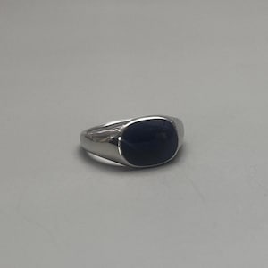 Puede incluir: Un anillo de sello plateado con una piedra rectangular azul oscuro. El anillo tiene una superficie pulida y reflectante y un diseño clásico y minimalista. La piedra está empotrada en la banda del anillo.