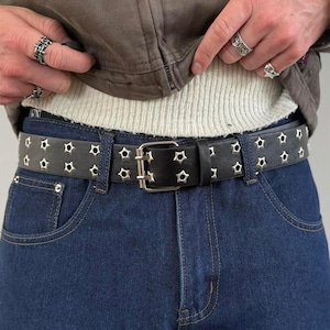 May include: Black leather belt with star-shaped cutouts and a silver buckle. The belt is worn with blue denim jeans. The belt has a double row of star-shaped cutouts. The buckle is silver and rectangular.