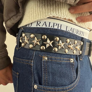 May include: Black leather belt with silver star and stud embellishments. The belt is worn with blue denim jeans and a white and black Ralph Lauren waistband. The belt has a silver buckle.