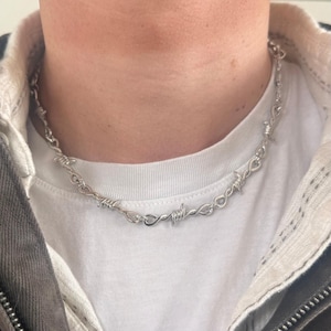 May include: A silver-coloured barbed wire necklace. The necklace is made of twisted metal and features sharp, pointed barbs. The necklace is worn around the neck and rests on a white t-shirt.