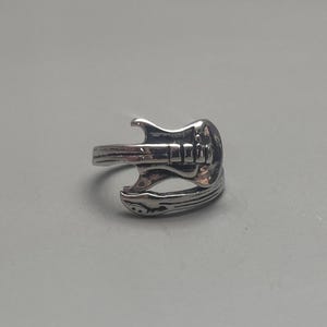 May include: A silver ring shaped like an electric guitar. The ring is detailed with the guitar's body, neck, and strings.