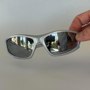 May include: Silver-framed sunglasses with dark lenses. The sporty eyewear features a wraparound design and a reflective surface. The sunglasses are held by a hand, showcasing the sleek, modern style.