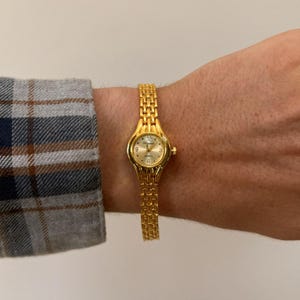 May include: A gold-toned wristwatch with a rectangular face and a linked band. The watch face displays the numbers 12, 3, 6, and 9. The watch is worn on a wrist, and the band is approximately 6 inches in length.