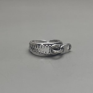 May include: A silver ring featuring a zipper design. The ring has a textured zipper pull and a smooth band.