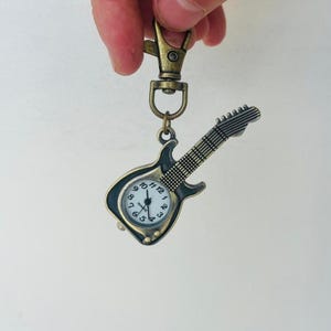 May include: A bronze-coloured guitar-shaped keyring with a clock face. The clock has black numbers and hands, and the guitar has a detailed fretboard and headstock. The keyring is attached to a bronze-coloured clasp.