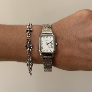 Puede incluir: Un reloj plateado con una esfera rectangular y una pulsera a juego. El reloj tiene números romanos y la palabra "NAIDU" impresa en la esfera. La pulsera presenta una serie de abalorios en forma de cruz.