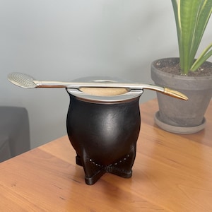 May include: A black leather-wrapped mate gourd with a silver-colored metal rim and base, featuring a matching silver-colored metal bombilla straw with a perforated filter end resting horizontally across the top opening of the gourd.