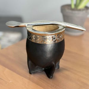 May include: A black mate gourd with a rounded base and a metallic rim featuring a repeating geometric pattern. A silver-colored metal bombilla straw with a perforated filter end rests horizontally across the top opening of the gourd.