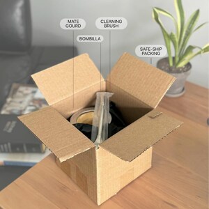 May include: An open brown cardboard shipping box containing a mate gourd, a metal bombilla, and a cleaning brush, with text labels pointing to each item and the safe-ship packing material inside the box.