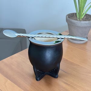 May include: A black mate gourd with a smooth, rounded body and a sturdy base, featuring a polished silver-colored metal rim. Resting across the top is a long, slender metal bombilla straw with a twisted handle and a perforated, oval-shaped filter.